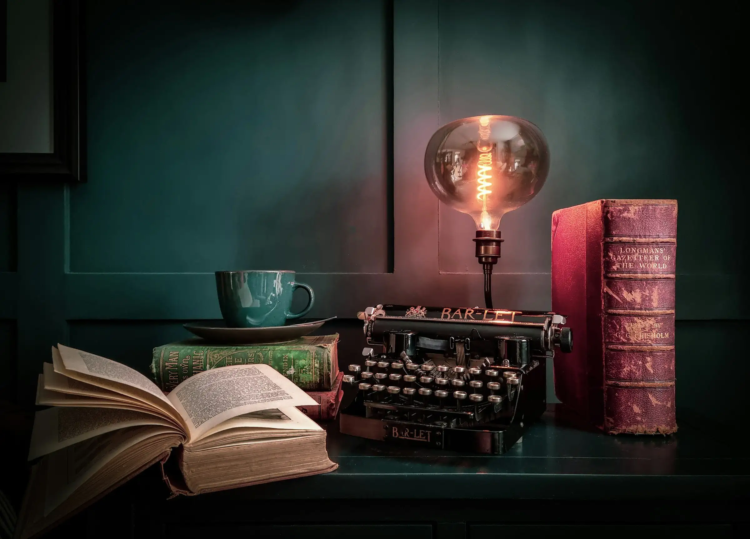 A dressed period desk with a typewriter, books and a glowing lamp